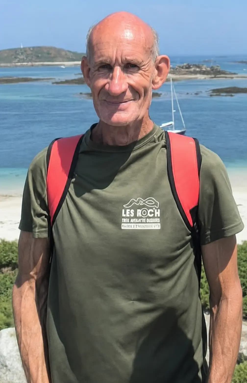 Denis devant une plage et des îlots, voilier au mouillage en arrière-plan